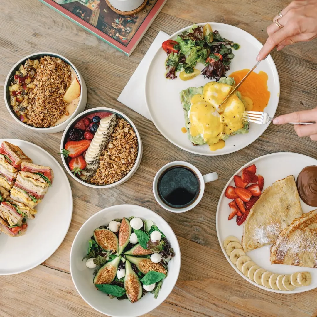 Alacati'nin en iyi kahvalti mekanlarindan birindeki zengin menüyü sergileyen bu ahşap masa görüntüsü, üzerinde yumurta benedict, çeşitli meyveli granola kaseleri, taze sandviçler, incirli salata, pudra şekerli krepler ve bir fincan kahve gibi lezzetli ve göz alıcı kahvalti çeşitleriyle dolu.