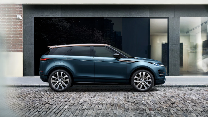 Koyu mavi renkte, çift renkli tavanı olan bir Range Rover Evoque model araç, şehirde park halinde duruyor. Arka planda modern bir binanın dış cephesi ve kaldırımda döşeli taşlar görünüyor.