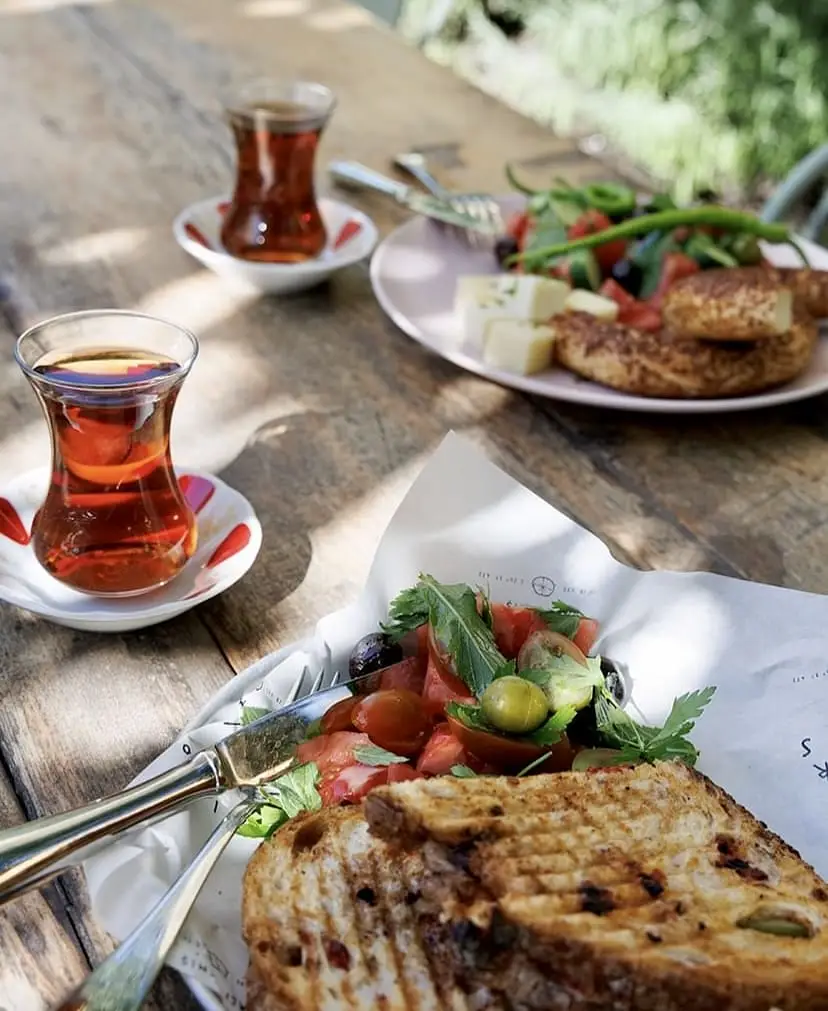 Alaçatı'nın en iyi kahvaltı mekanlarından birinde, ahşap bir masada servis edilen, ızgara tost, taze domates, zeytin ve yeşilliklerle dolu bir salata, yanında iki bardak demli çay ve peynir, simit ile çeşitli sebzelerden oluşan zengin bir Türk kahvaltısı tabağı, güneşin altında davetkar bir atmosfer sunuyor.