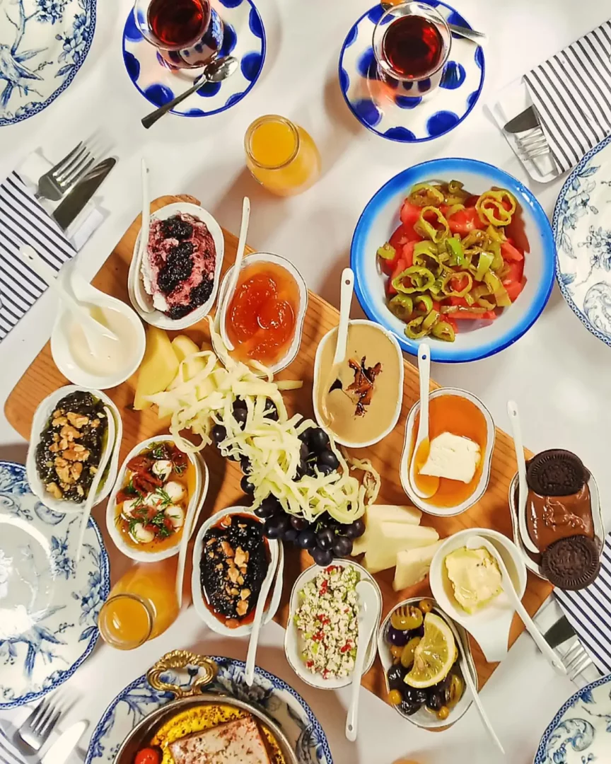 Alaçatı'nın en iyi kahvaltı mekanlarından birinde sunulan zengin bir Türk kahvaltısı masası, ahşap servis tahtasında çeşitli reçeller, ballı kaymak, tahin, farklı peynir çeşitleri, zeytinler, sarma, taze sebzeler, üzüm, Oreo bisküvili çikolata ve diğer birçok lezzetle dolu küçük beyaz kaselerin yanı sıra iki bardak çay ve portakal suyuyla cömertçe donatılmış bir sofrayı gösteriyor.