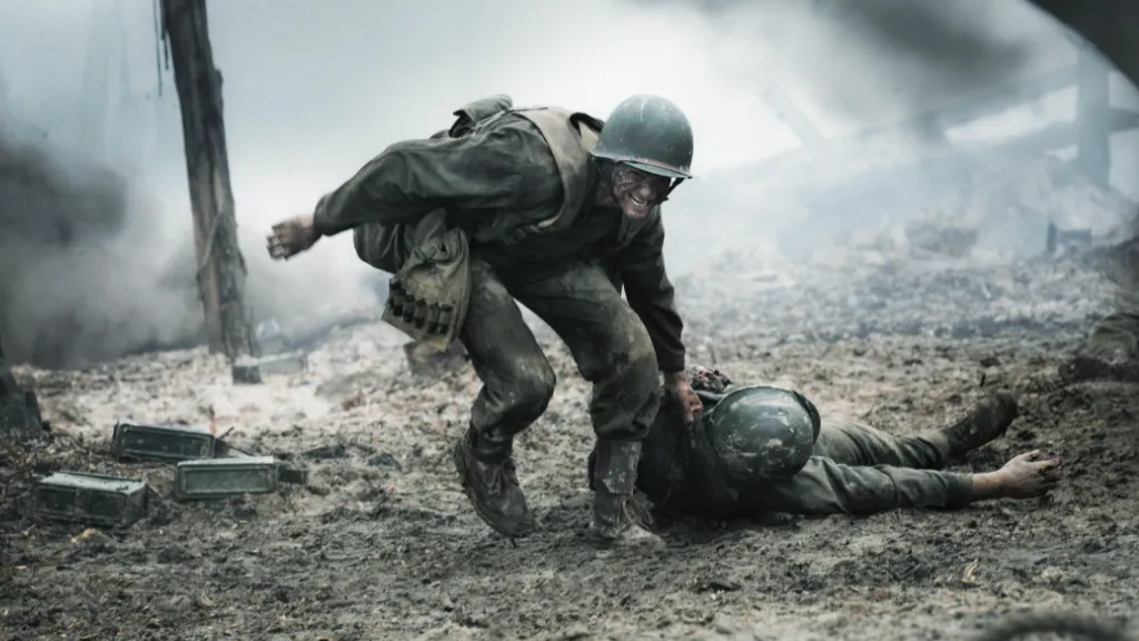Savaş Vadisi (Hacksaw Ridge) filminden bir sahne: Askerî üniformalı bir adam, savaş alanında yerde yatan yaralı bir askeri kurtarmaya çalışıyor. Dumanlar, molozlar ve çamurlu zemin savaşın şiddetini yansıtıyor.