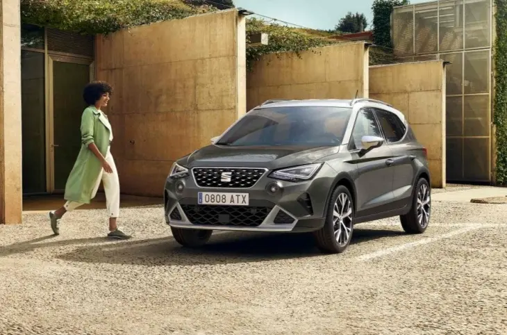Gri renkte bir Seat Arona model araba, modern bir binanın önünde park halinde duruyor ve yanında yürüyen bir kadın görünüyor.