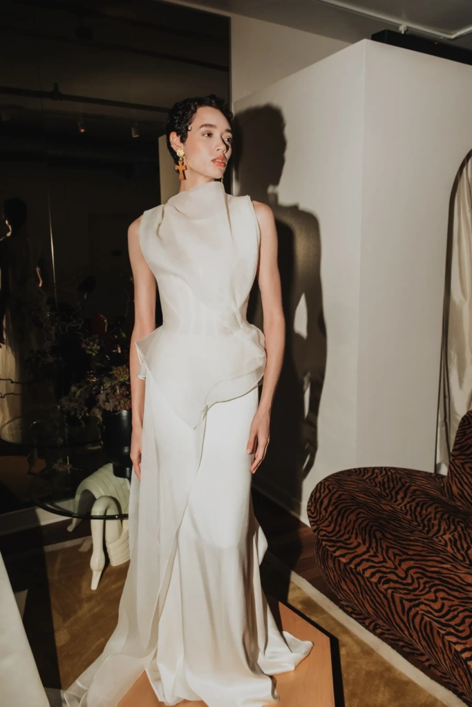 Modern mimariden ilham alan bu zarif gelinlik, heykelsi formları ve asimetrik drapeleriyle öne çıkıyor. Sept Bridal imzalı tasarım; sade ama etkileyici bir duruş arayan gelinler için sofistike, sanatsal ve zamansız bir seçenek sunuyor.