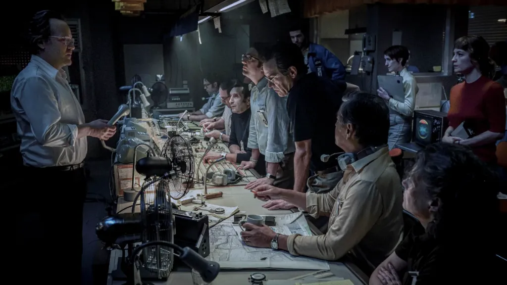 % Eylül filminden bir sahne: Kontrol odasında yoğun bir toplantı: Eski tip ekipmanlarla dolu bir NASA tarzı kontrol merkezinde, çoğunluğu erkeklerden oluşan bir grup görevli ciddi bir şekilde görev başında. Bir adam elindeki mikrofonla konuşurken diğerleri ekranlara, kağıtlara ve önlerindeki cihazlara odaklanmış durumda. Ortam karanlık ve yoğun; acil bir durum izlenimi veriyor. Retro teknolojiler ve kıyafetler, sahnenin 1960–70'lerde geçtiğini ima ediyor.