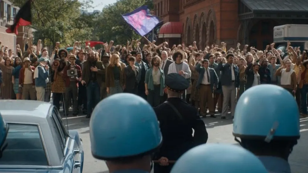Şikago Yedilisi'nin Yargılanması (The Trial of the Chicago 7) filminden bir sahne: Ellerini havaya kaldırmış büyük bir protestocu kalabalık, polis barikatının karşısında duruyor. Ön planda mavi miğferli polisler ve bir polis aracı görülürken, arka planda öfkeli kalabalık slogan atıyor ve pankartlar taşıyor. Sahne, toplumsal hareketlerin baskıyla karşılaştığı gerilimli atmosferi yansıtıyor.