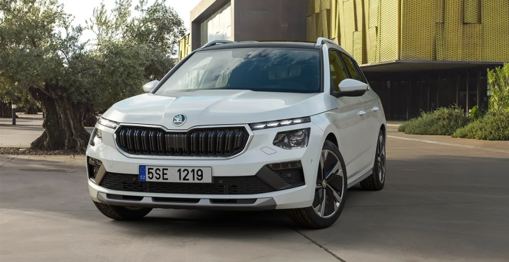 Beyaz renkte bir Skoda Kamiq model araba, modern bir binanın ve ağaçların önünde duruyor. Aracın ön farları, ızgarası ve dinamik tasarımı belirgin bir şekilde görülüyor.