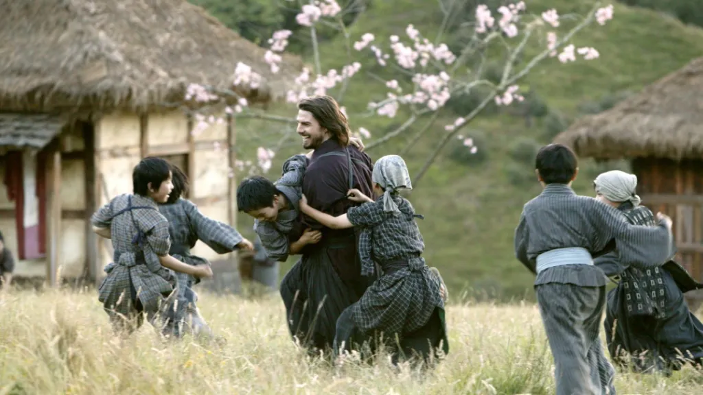 Son Samuray (The Last Samurai) filminden bir sahne: Tom Cruise’un canlandırdığı karakter, geleneksel Japon kıyafetleri giymiş çocuklarla birlikte çiçek açmış ağaçların arasında neşeyle oyun oynuyor. Arka planda geleneksel Japon kulübeleri görülüyor, sahne huzur ve mutluluk atmosferi yansıtıyor.