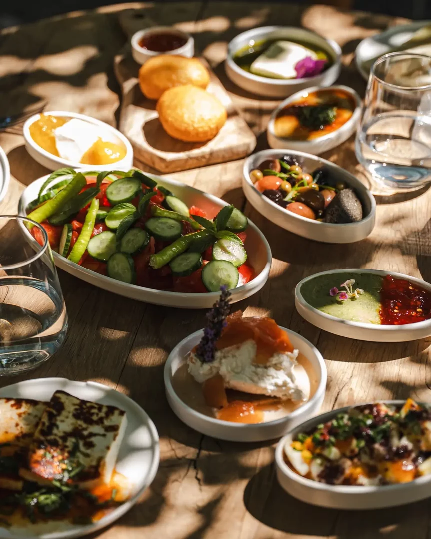 Alacati'nin en iyi kahvalti mekanlarindan birinde, güneşin vurduğu ahşap masada, taze salata, pişi, kızarmış hellim, zeytin, peynir ve reçel gibi yöresel lezzetlerle dolu zengin bir Türk kahvaltısı masası.