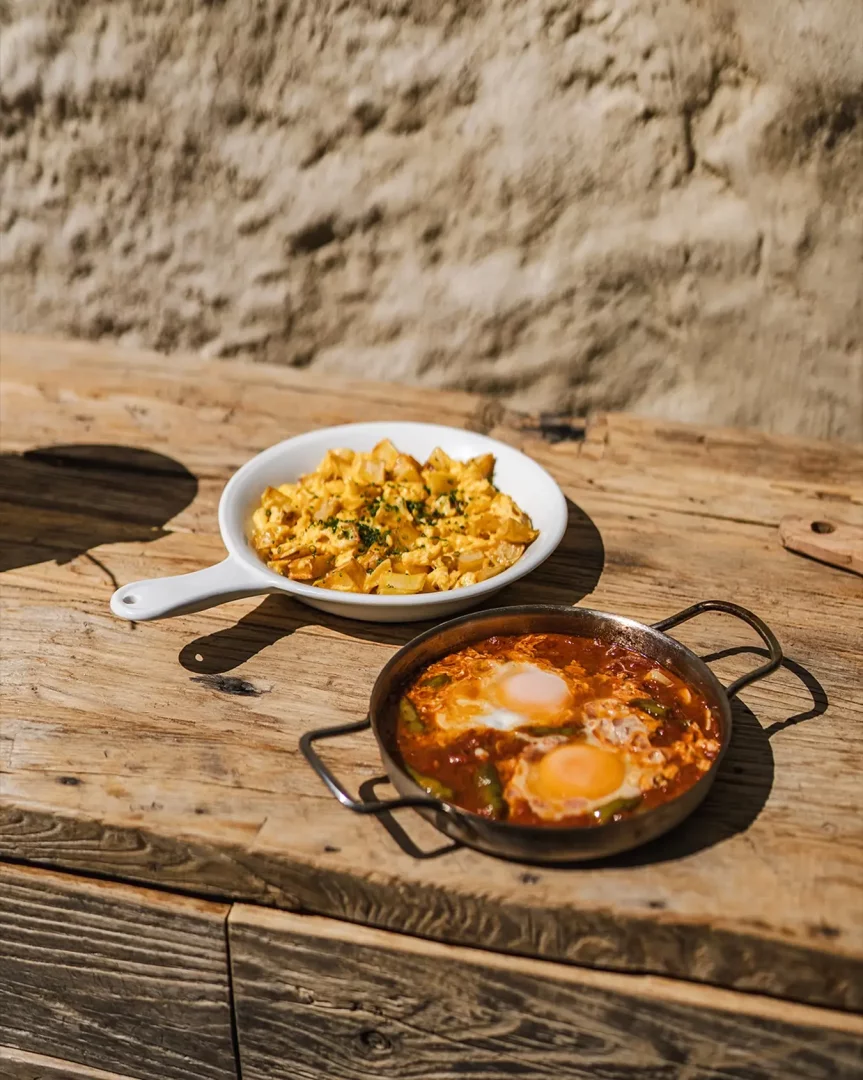 Alaçatı'nın en iyi kahvaltı mekanlarından birinde, rustik ahşap bir masada, domates soslu iki göz yumurta içeren menemen tavası ile patatesli omletin olduğu beyaz sahan tabağı güneş ışığında iştah açıcı görünüyor.