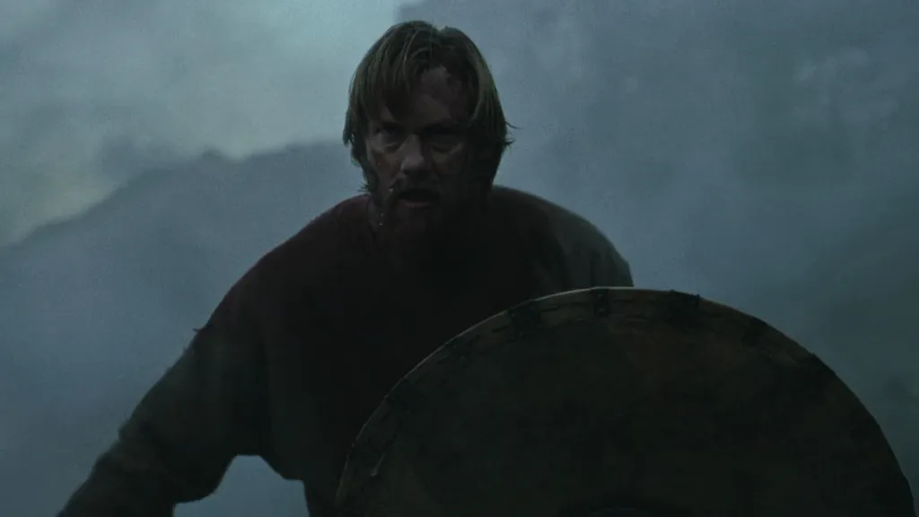 The Northman filminden bir sahnede, sisli bir savaş alanında elinde yuvarlak ahşap kalkan tutan, kanlar içindeki Viking savaşçısı kararlı bir ifadeyle ileriye bakıyor. Arka planda bulanık dağ manzarası yer alıyor.