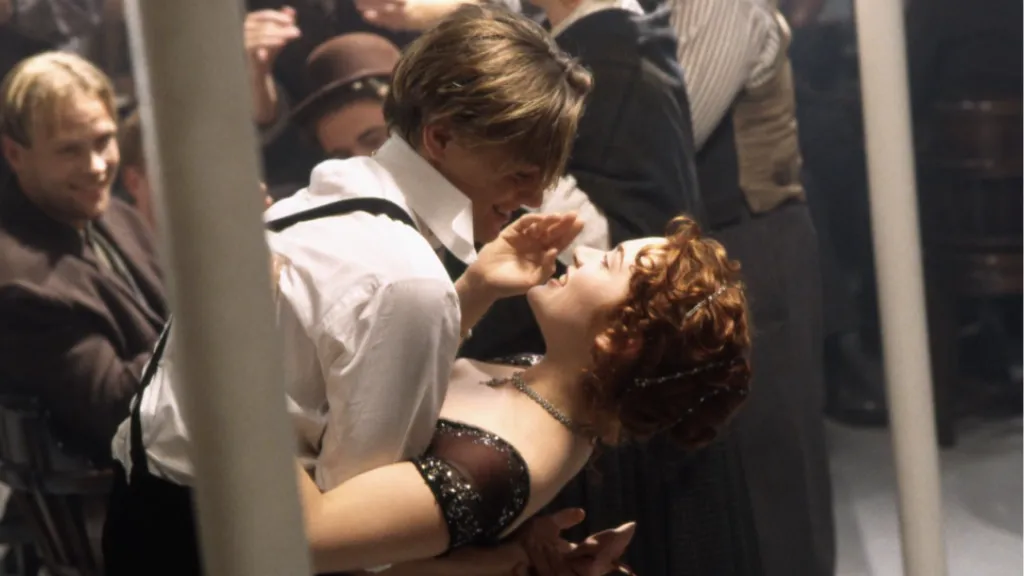 Titanic filminden ikonik bir sahne: Jack ve Rose, geminin alt kat salonunda dans ederken kahkahalarla eğleniyor. Jack, Rose’u geriye doğru eğmiş, ikisi de göz göze bakıyor. Etrafları neşeyle dolu kalabalık bir ortam.