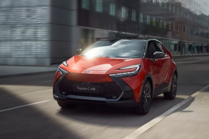 Kırmızı ve siyah renkte, çift renkli bir Toyota C-HR model araç, şehirde yolda ilerlerken çekilmiş. Aracın dinamik tasarımı ve far detayları ön planda, arka planda ise modern binalar görülüyor.