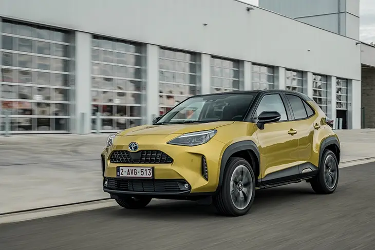 Açık sarı renkte, çift renkli tavanı olan bir Toyota Yaris Cross model araba, modern bir binanın önünde yolda ilerliyor.