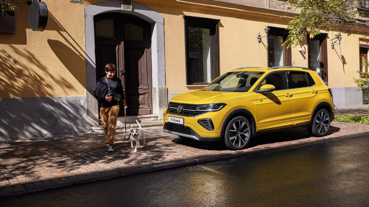 Sarı renkte bir VW T-Cross model araba, eski bir binanın önünde bir kaldırımda duruyor. Arabanın yanında, tasma ile bir köpekle yürüyen bir adam görünüyor.