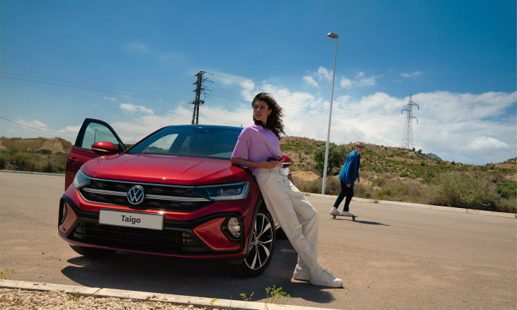 Kırmızı renkte bir VW Taigo model araba, çöl benzeri bir yolun kenarında duruyor. Arabaya yaslanmış, elinde telefon olan bir kadın ve arka planda kaykay yapan genç bir adam görünüyor.