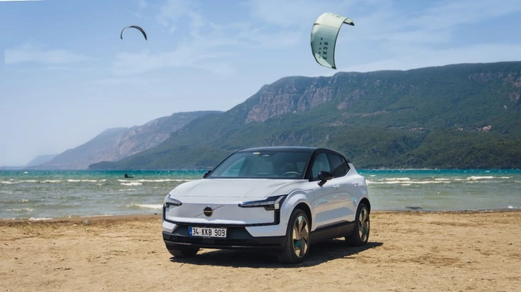 Beyaz renkli Volvo EX30 model elektrikli SUV araba, arka planda deniz ve dağ manzarası olan kumsalda duruyor. Gökyüzünde iki adet uçurtma sörfü (kitesurf) yapan kişi görünüyor.