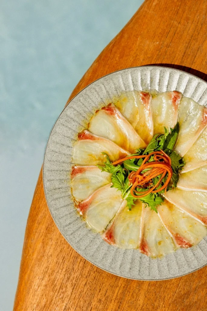 Bodrum'un en iyi restoranlarının sunduğu zengin lezzetleri temsil eden, yuzu trüf soslu ince dilimlenmiş akya sashimi tabağı, taze yeşillikler ve portakal rengi rendelenmiş havuçlarla süslenmiş olarak şık bir ahşap masa üzerinde yer alıyor.