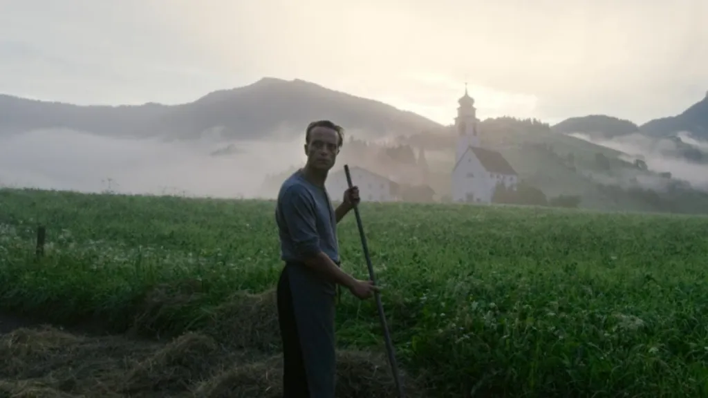 A Hidden Life filminden pastoral bir sahne: Sabah sisleriyle örtülü dağların eteklerinde uzanan yeşil bir tarlada, elinde tırmık tutan bir adam duruyor. Arka planda beyaz bir kilise ve dağ silueti görülüyor. Sahne, doğa içinde geçen sade ve dingin bir yaşamı yansıtıyor.