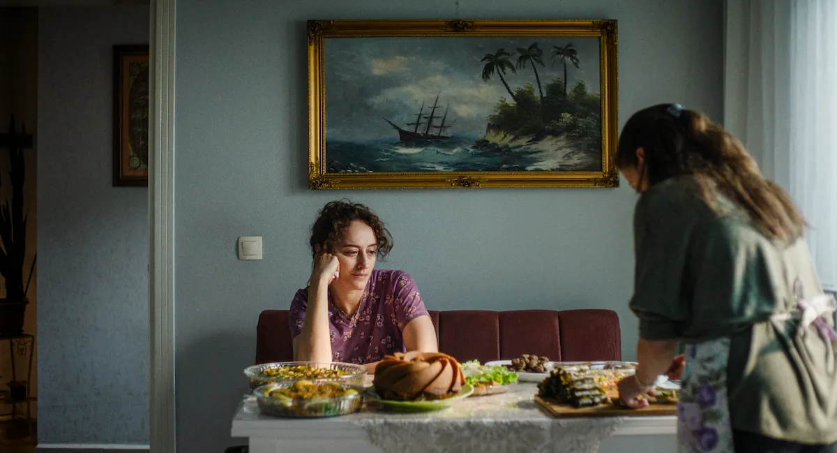 İstanbul Film Festivali: Altın Lale Yarışması filmlerinden Yalnız'ın bu etkileyici film karesinde, Selin Tezcan yemeklerle dolu bir masada düşünceli bir şekilde otururken, arkasında bir kadın yemekleri hazırlıyor ve duvarda fırtınalı bir deniz tablosu asılı.
