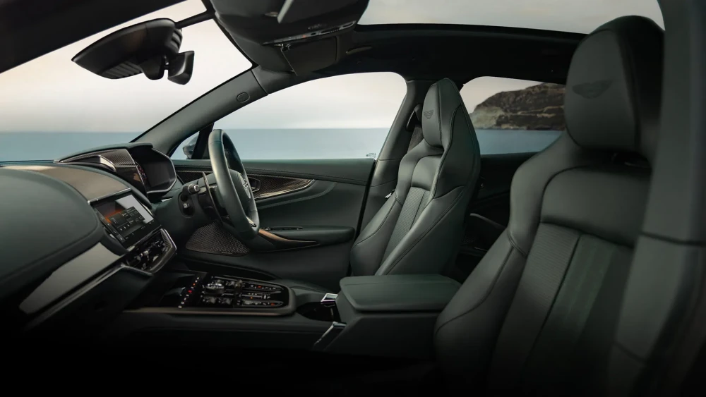 Aston Martin DBX 707 lüks SUV'nin yolcu tarafından çekilmiş bu detaylı iç mekan tasarımında, koyu renk delikli deri koltuklar, karbon fiber kaplamalı kokpit, direksiyon simidi ve orta konsol ile birlikte dışarıda görünen huzurlu deniz manzarası bütünleşiyor.