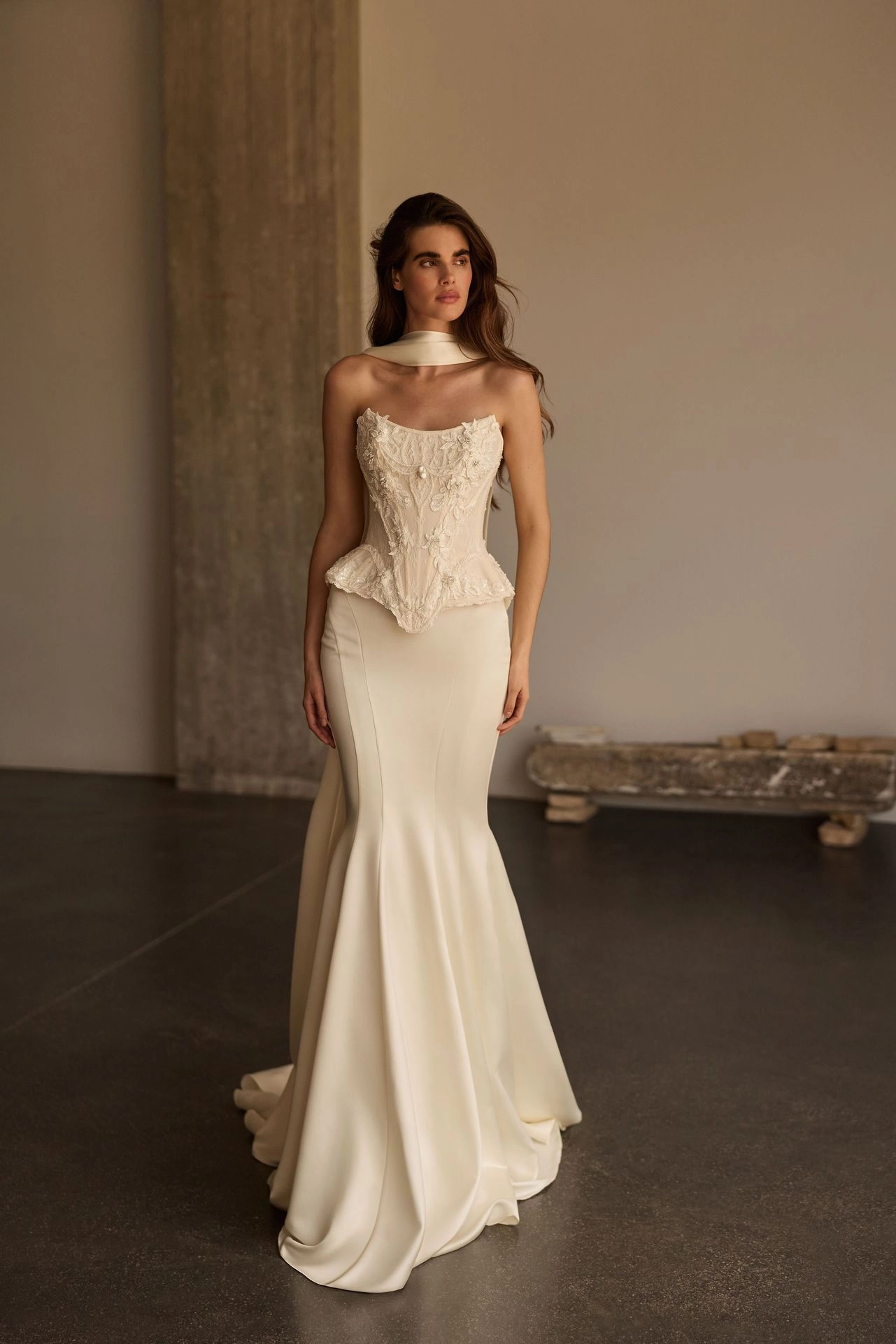 Bu şık balık gelinlik modelinde, incelikle işlenmiş dantel ve peplum detaylı straplez korse büstiyer, ipek saten etekle birleşerek vücut hatlarını kusursuzca vurgularken, tamamlayıcı boyun bandı modern ve iddialı bir zarafet sunuyor.