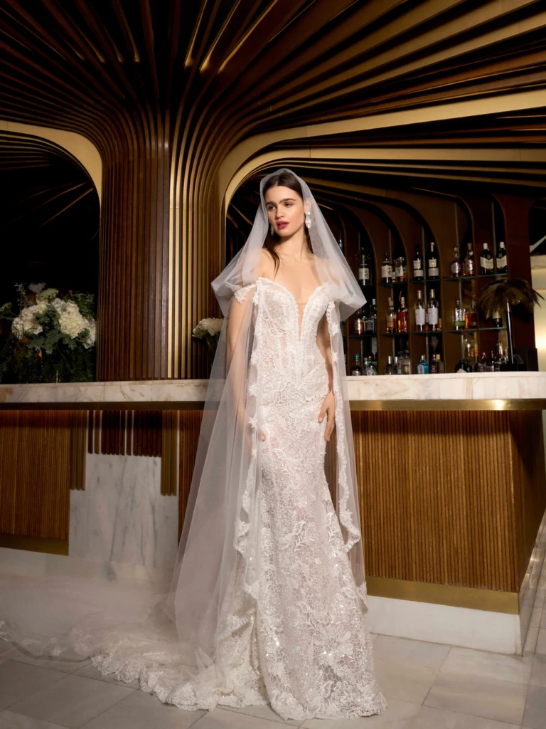 Modern ve şık bir otel barının önünde duran, Galia Lahav imzalı, omuzları açıkta bırakan, derin V yakalı, zarif dantel işlemelerle süslenmiş bembeyaz balık kesim gelinliği ve yere kadar uzanan tül duvağıyla çekici bir gelin.