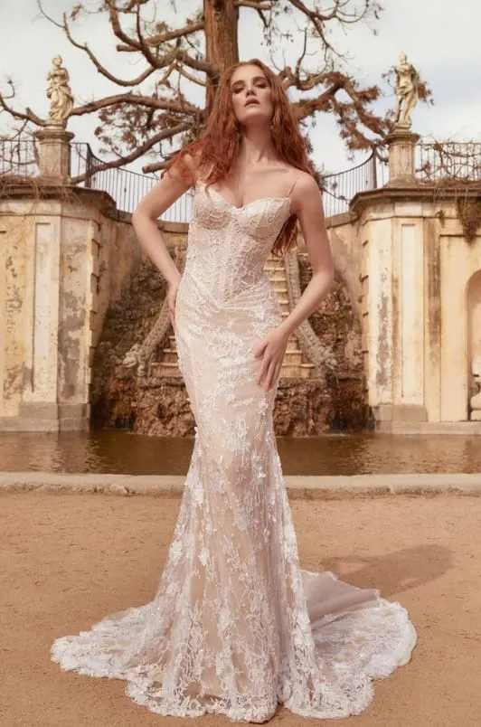 Kızıl saçlı bir model, zarif bir duruşla elleri belinde poz verirken, Galia Lahav'ın imzasını taşıyan, tümüyle işlemeli ve ışıltılı straplez beyaz bir balik gelinlik giymiş; arkasında detaylı işlenmiş tarihi bir taş yapı görünüyor.