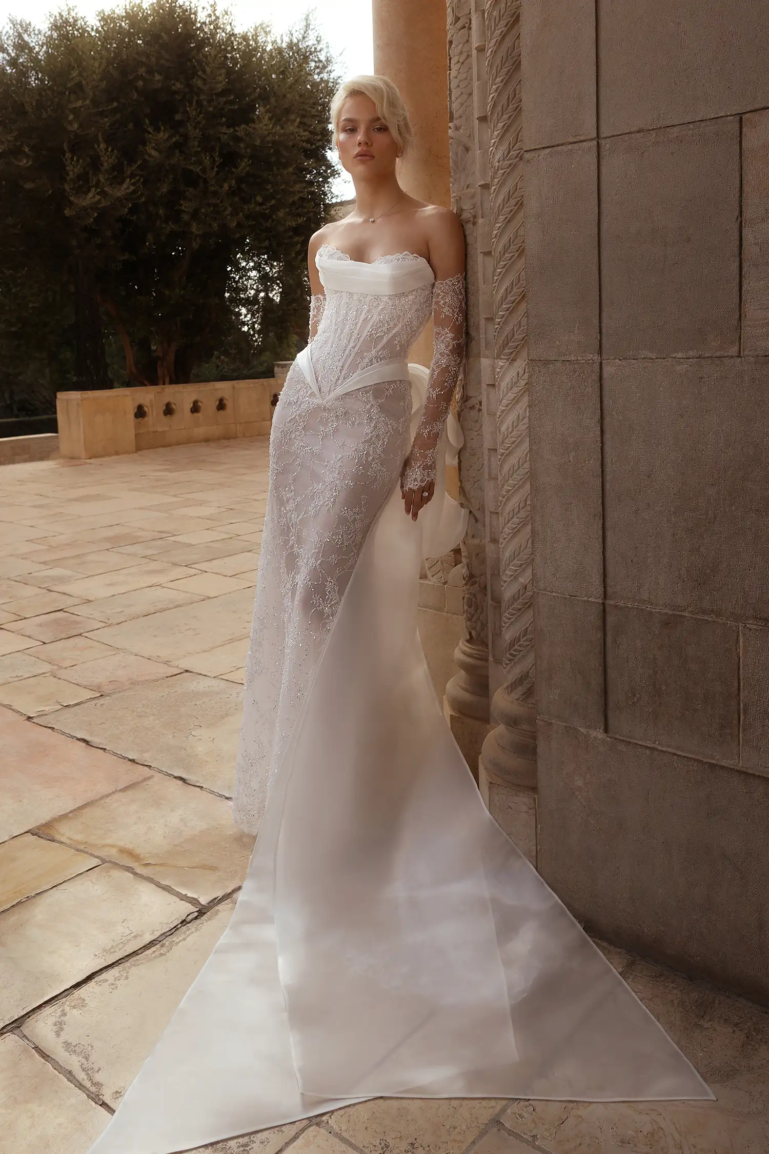 Pnina Tornai straplez kalp yaka, gövdesi dikey şeritli transparan korse formunda, dantel eldiven detaylı ve arkadan eklenen saten pelerinli ışıltılı dantel işlemeli balık model gelinlik.