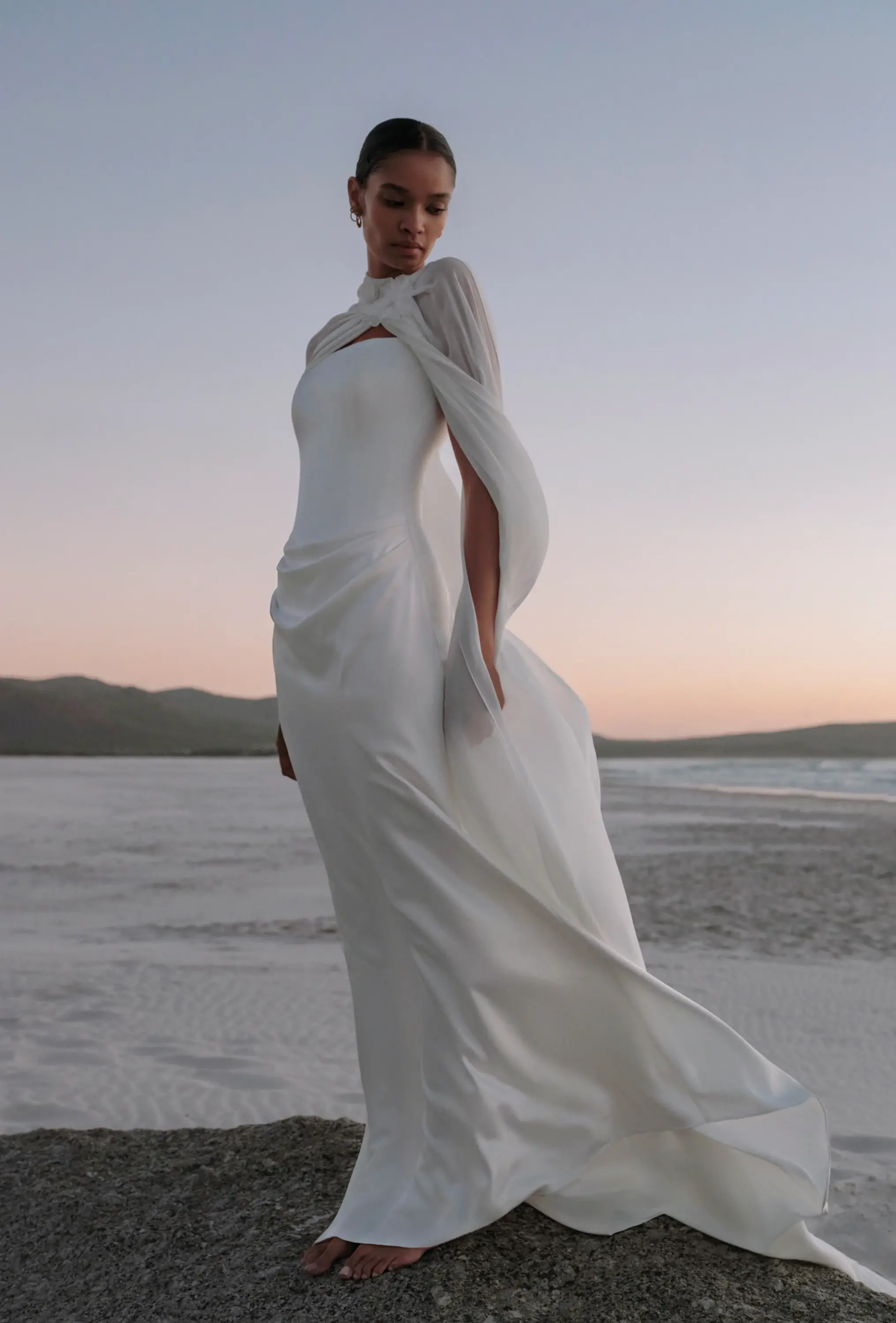 Project Bridal straplez yaka, sade krep kumaştan vücuda oturan, kalça kısmında minimal drape detaylı ve omuzlardan dökülen yüksek yakalı, şifon pelerin aksesuarlı balık model gelinlik.
