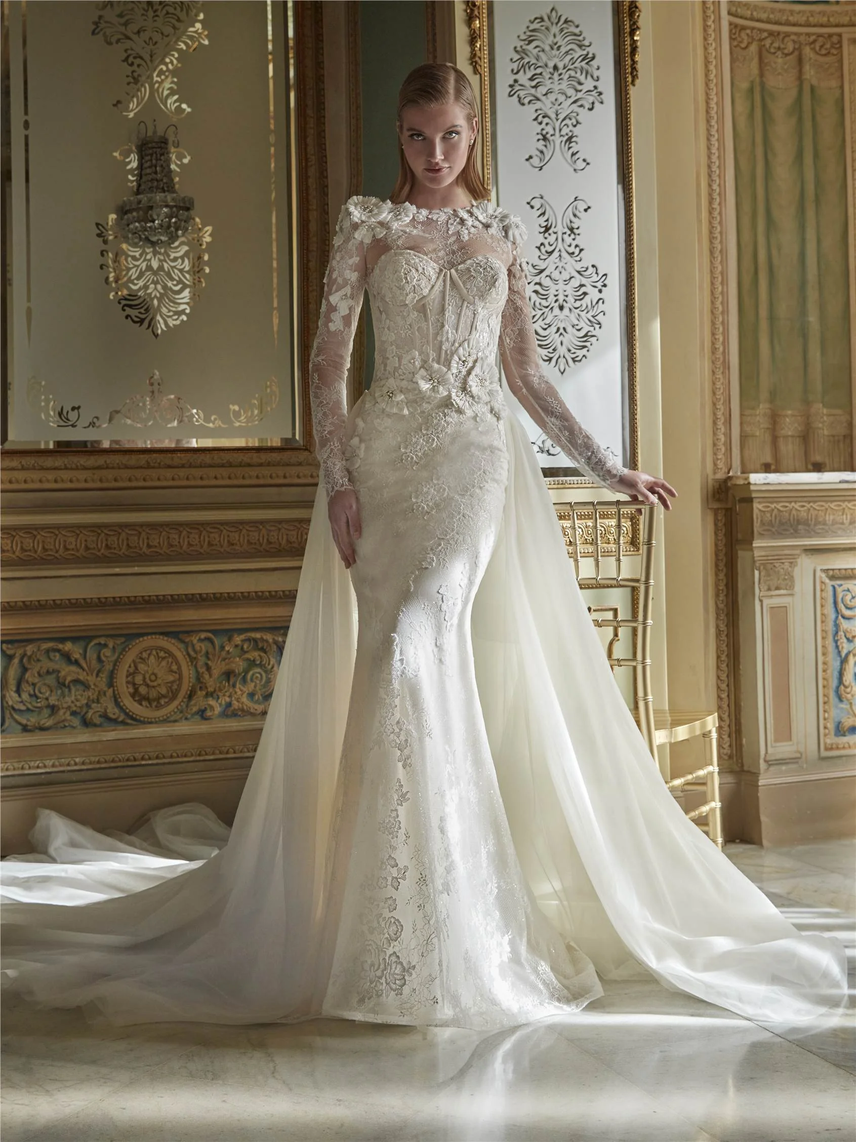Pronovias Priveé illüzyon yaka detaylı, uzun dantel kollu, omuzları ve beli 3D çiçek aplikeleriyle bezeli, gövdesi dikey şeritli korse formunda ve arkadan eklenen görkemli tül pelerinli balık model gelinlik.