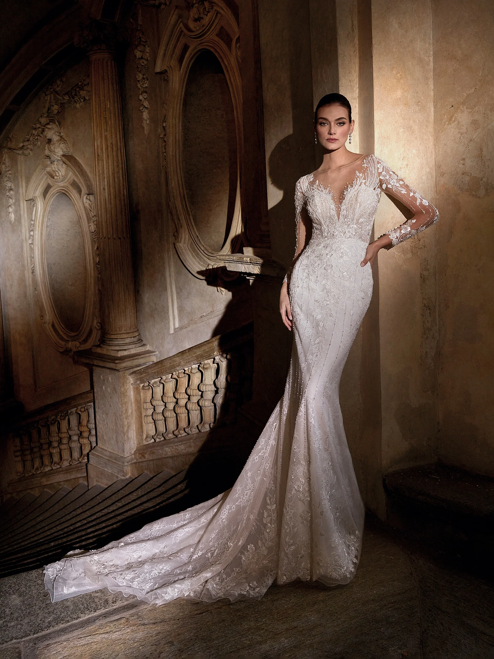 Pronovias Priveé illüzyon yaka ve derin dekolte efektli, uzun kollu, omuzlardan eteğe doğru dikey ışıltılı hatlarla inen ve kristal işlemeli dantel aplikelerle zenginleştirilmiş balık model gelinlik.