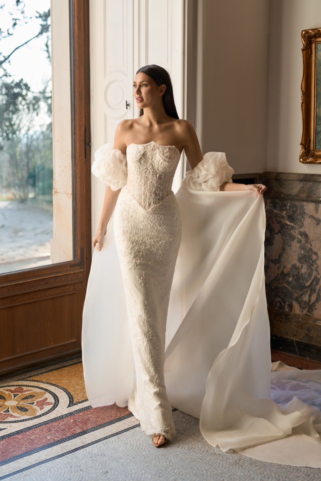 Sima Couture straplez kalp yaka, gövdesi yoğun inci ve kristal işlemeli korse büstiyer formunda, kollarda hacimli tül fırfır detayları bulunan ve arkadan eklenen uzun tül pelerinli balık model gelinlik.