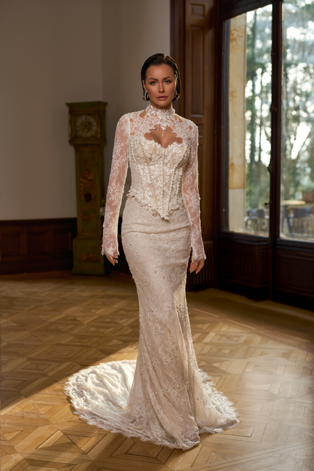 Sima Couture yüksek dantel yakalı, pencereli dekolte detaylı ve uzun dantel kollu; gövdesi dikey inci dizileri ve kristal taşlarla yoğun şekilde işlenmiş korse büstiyer formunda, tamamı floral dantel kaplı balık model gelinlik.