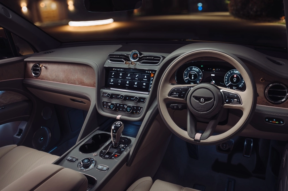 Bentley Bentayga lüks SUV'un gece çekilen iç mekan görüntüsünde, ahşap detaylarla zenginleştirilmiş ön konsol tasarımı, geniş dokunmatik ekran, dijital gösterge paneli ve çok fonksiyonlu direksiyonun bej ve koyu gri renk uyumuyla modern ve sofistike bir atmosfer sunuluyor.