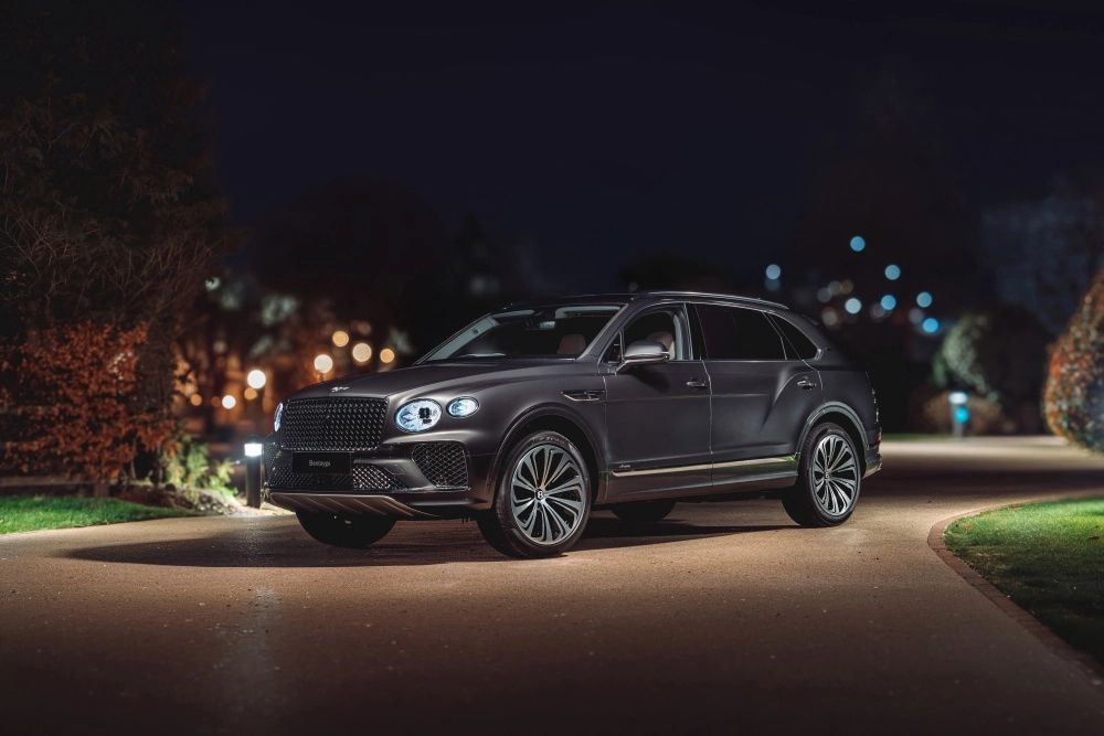 Karanlık bir gecede, lüks SUV sınıfının göz alıcı mat gri Bentley Bentayga modeli, güçlü ön tasarımı ve yanan farlarıyla aydınlatılmış bir yolda park halinde duruyor, arka planda bulanık ağaçlar ve parlayan ışıklar görünüyor.