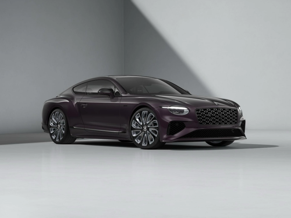 Açık gri minimalist bir stüdyo ortamında, ön ve sol çaprazdan görünen, koyu mor renkli, aerodinamik hatlara sahip Bentley Continental GT plug-in hibrit spor coupé modelinin lüks ve modern dış tasarımı vurgulanıyor; parlak jantları ve özgün ön ızgarası dikkat çekiyor.