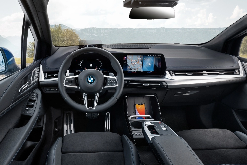 Geniş açılı bu fotoğraf, BMW 230e xDrive Active Tourer şarj edilebilir hibrit modelinin modern iç mekan tasarımını gözler önüne seriyor; kavisli dijital ekran, spor direksiyon simidi ve orta konsolda kablosuz şarj ünitesinde duran akıllı telefon ile teknolojik ve aile dostu bir atmosfer sunuyor.
