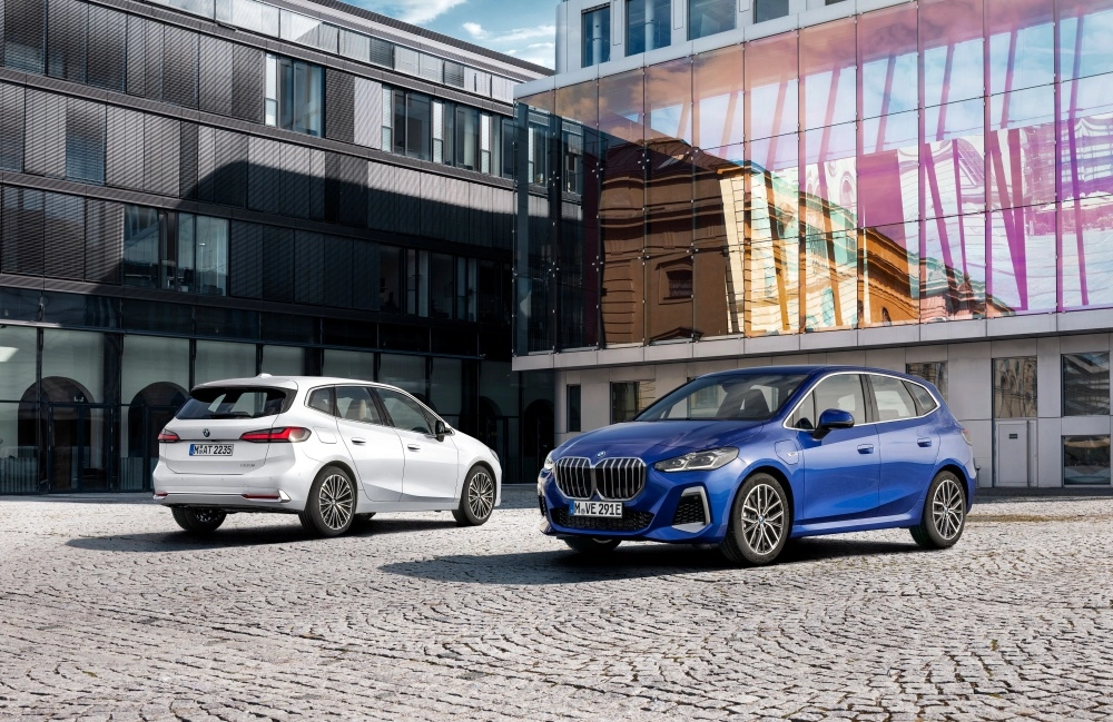 Ortak noktaları olan ancak farklı renklerdeki iki BMW 2 Serisi Active Tourer şarj edilebilir hibrit aile otomobili, modern binaların önündeki taş döşeli yolda duruyor; mavi olan öne, beyaz olan ise arkaya dönük.