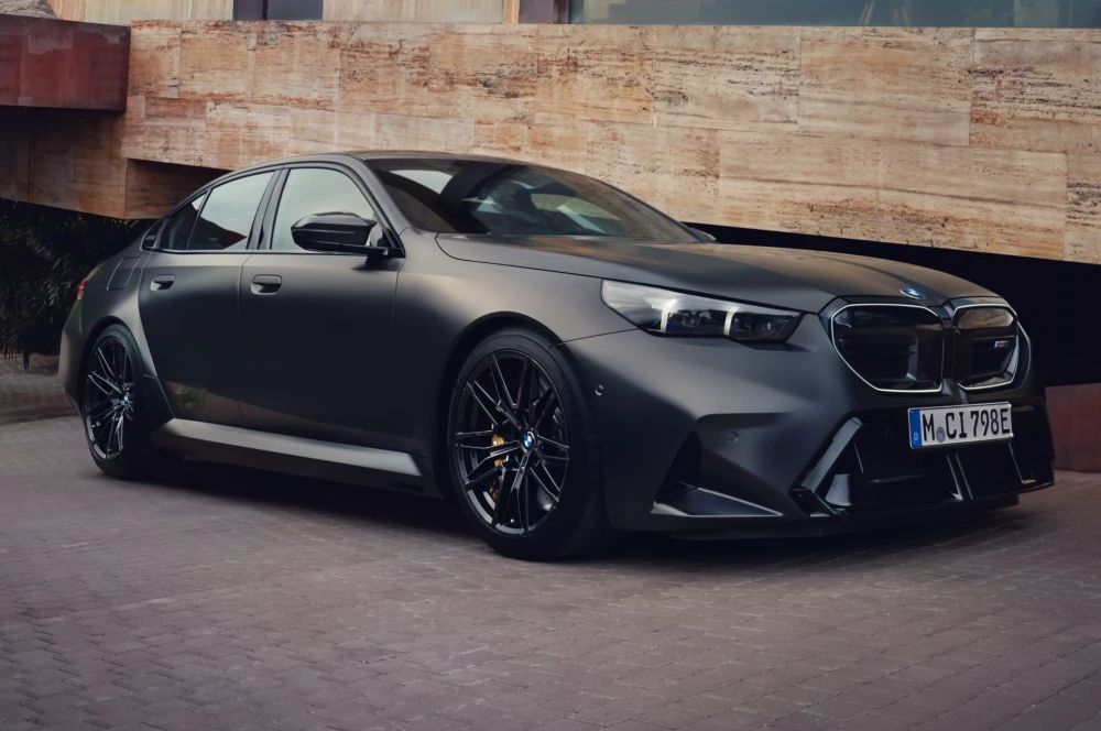 Mat siyah renkli, modern ve agresif hatlara sahip yeni nesil bir BMW M5 sedan, tuğla döşeli bir zeminde park edilmiş halde, aracın dinamik dış tasarımı ve şarj edilebilir hibrit süper sedan kimliği öne çıkarılıyor.