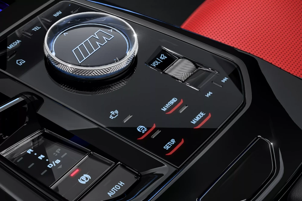 BMW M5 sedanın şık iç mekan tasarımından bir kesit gösteren bu görselde, parlak siyah orta konsolun merkezinde M logosuyla vurgulanmış iDrive kumandası, M Hybrid ve M Mode tuşları ile vites seçici belirgin şekilde yer alıyor.