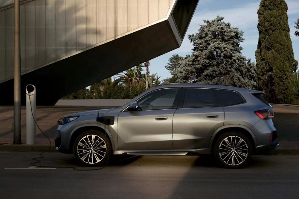 Modern bir binanın ve yeşilliklerin önünde park halinde duran, gümüş gri rengindeki yeni BMW X1 xDrive25e plug-in şarj edilebilir hibrit SUV otomobil, sol ön çamurluğundaki porttan bir şarj istasyonuna bağlı olarak dış tasarımıyla göz alıyor.