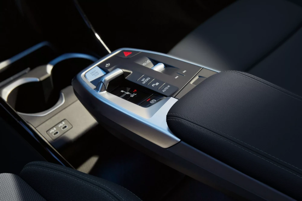 BMW X1 xDrive25e plug-in şarj edilebilir hibrit SUV otomobilin zarif iç mekan tasarımını vurgulayan, ergonomik iDrive kumandası, kompakt vites seçici, multimedya kontrolleri, Start/Stop ve Auto H düğmelerini içeren modern orta konsolun detaylı görünümü, solunda bardaklık ve USB-C girişleriyle tamamlanıyor.