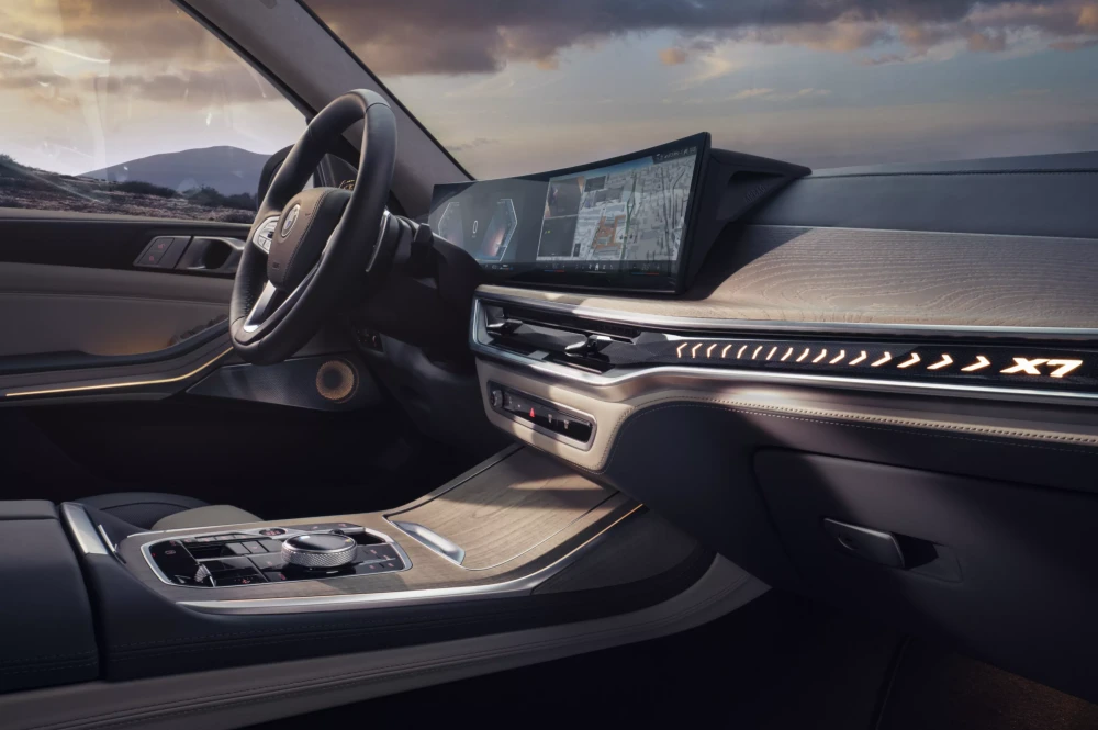 BMW X7 lüks aile SUV'unun modern iç mekan tasarımı, büyük kavisli dijital ekranı, zarif ahşap kaplamaları ve yolcu tarafındaki ışıklı X7 logosuyla ön konsolunu vurguluyor, lüks ve teknoloji dolu bir sürüş deneyimi sunuyor.
