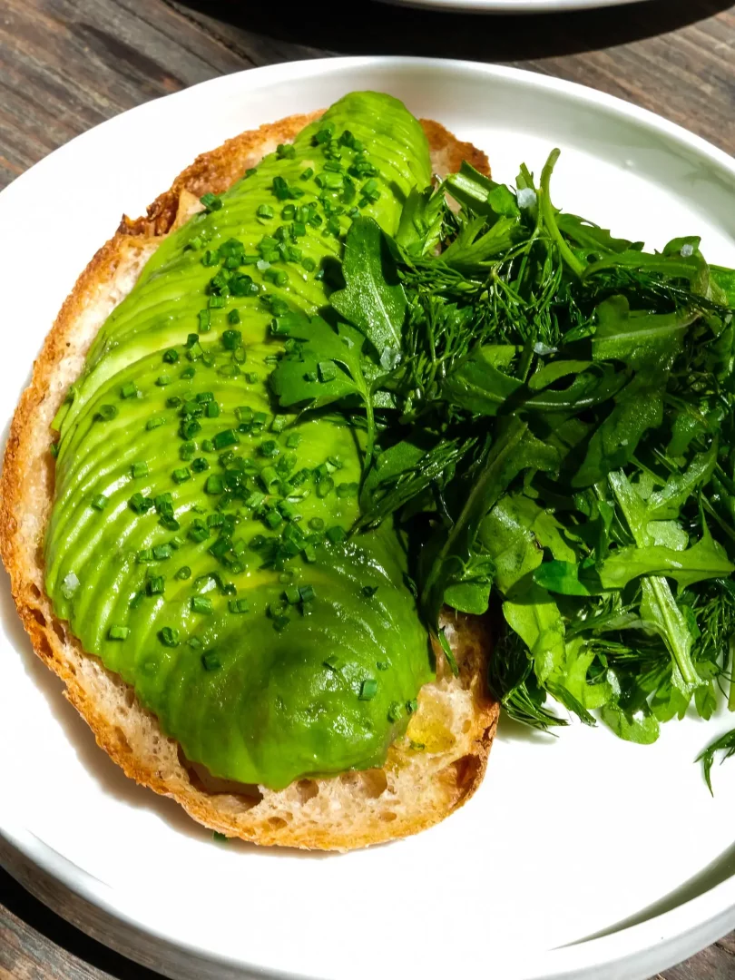 Alaçatı'nın en iyi kahvaltı mekanlarında sunulan bu iştah açıcı karede, çıtır kızarmış ekmek üzerinde özenle dizilmiş taze avokado dilimleri ve frenk soğanı serpilmiş, yanında bol yeşillikli bir salata ile nefis bir kahvaltı tabağı güneşli bir masa üzerinde sergileniyor.