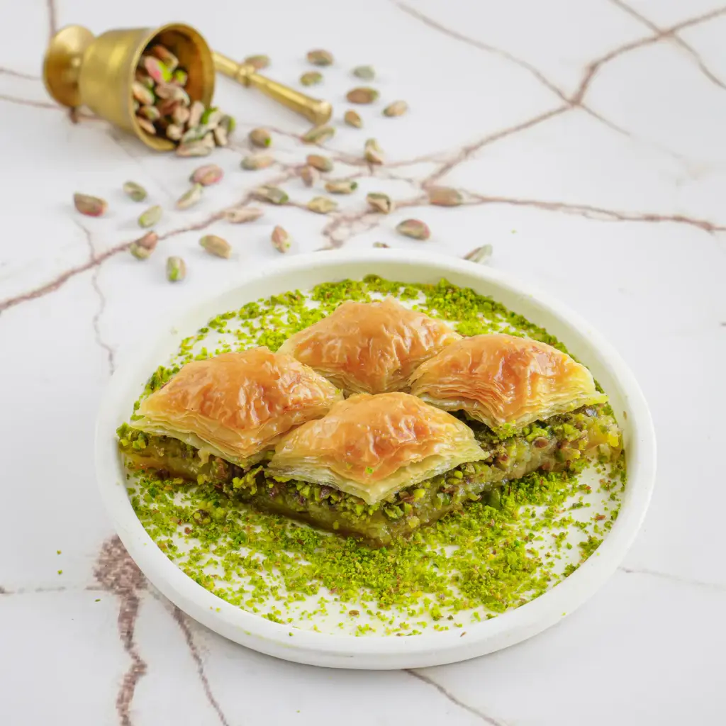 Beyaz mermer zemin üzerinde sunulan klasik fıstıklı baklava. Bolca serpilmiş yeşil toz fıstıkla kaplı beyaz tabakta dört dilim altın sarısı çıtır baklava yer alıyor. Arka planda devrilmiş pirinç bir havandan bütün Antep fıstıkları etrafa