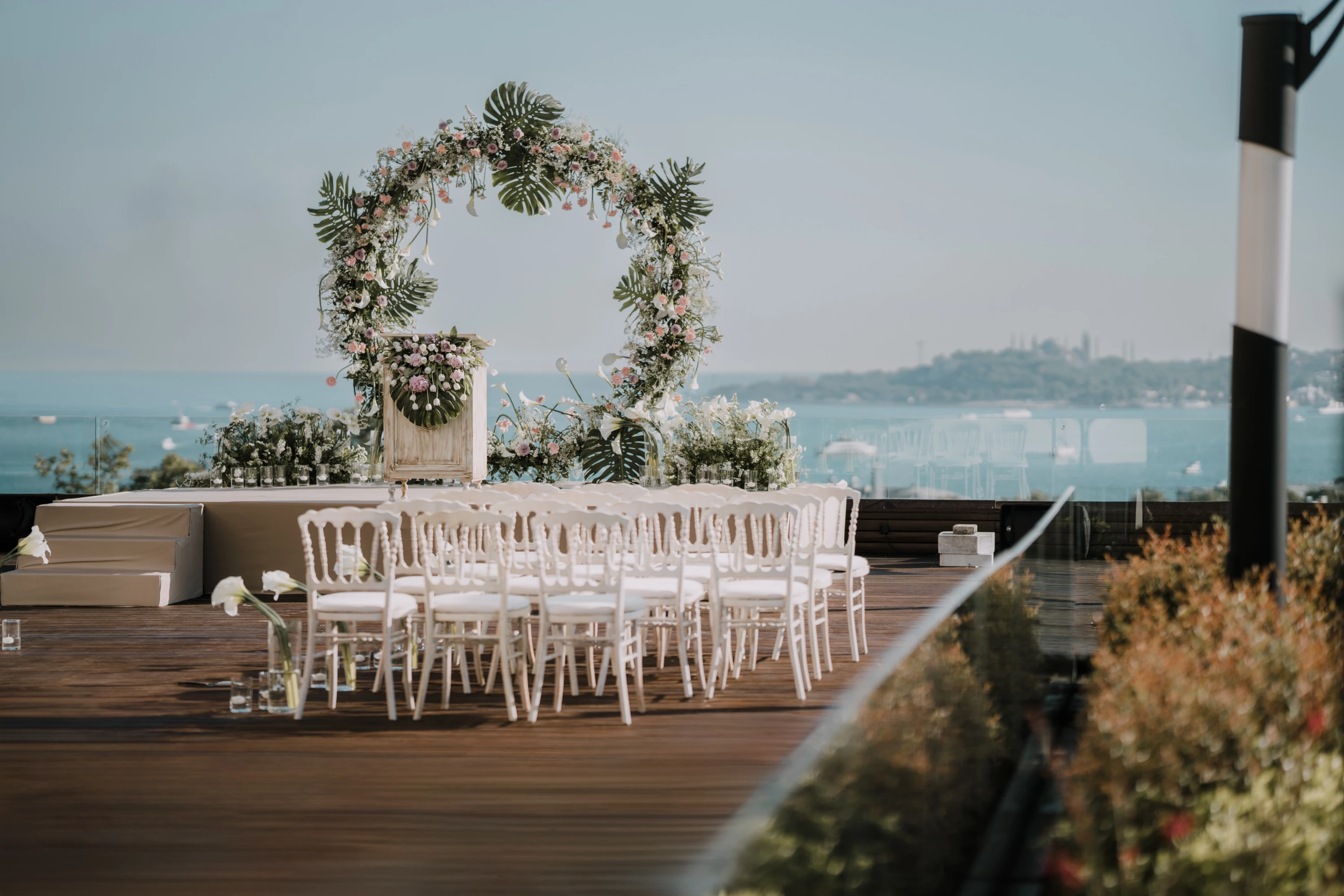 Conrad İstanbul'un Boğaz manzaralı terasında, çiçeklerle bezenmiş yuvarlak bir kemer ve önündeki sahneye doğru sıralanmış beyaz sandalyelerle, uzakta İstanbul'un tarihi siluetinin göründüğü bir düğün töreni veya özel etkinlik için hazırlanmış zarif bir açık hava düzenlemesi.