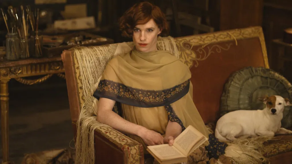 The Danish Girl (Danimarkalı Kız) filminden bir sahne: Şık bir odada, vintage bir koltukta oturan zarif görünümlü bir kişi elinde açık bir kitap tutuyor. Omzunda nakışlı ince bir şal olan kişi doğrudan kameraya bakarken, yanında koltuğa uzanmış beyaz ve kahverengi renkli küçük bir köpek yer alıyor. Arka planda sanat malzemeleri ve fırçalarla dolu eski bir masa dikkat çekiyor. Atmosfer, 1920’ler Avrupa'sının sanatsal ve sofistike havasını yansıtıyor.