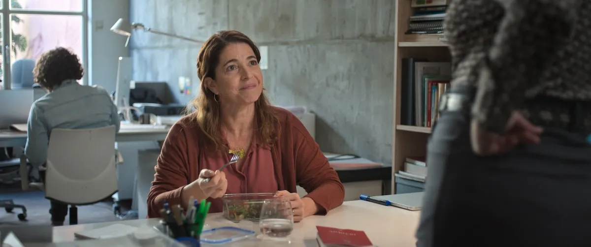 İstanbul Film Festivali: Arkadaşım Eva filminden bir karede, orta yaşlı bir kadın modern ofis masasında oturmuş, yemek kabından salata yiyip hafifçe gülümseyerek karşısındakiyle konuşur gibi yukarı bakıyor; arka planda bulanık şekilde başka bir çalışan da görülüyor.