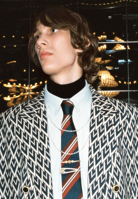 Lüks bir ortamda poz veren genç erkek model, V-logolu kravat iğnesi ve inci kolye gibi modern aksesuarlarla tamamladığı geometrik desenli blazer ceket, balıkçı yaka ve çizgili kravat kombinasyonuyla ünlü erkek giyim markalarının zamansız ve yenilikçi stilini gözler önüne seriyor.