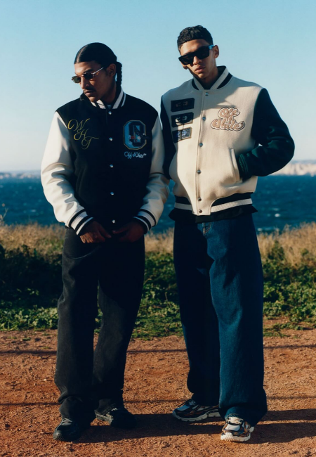 Mavi gökyüzü ve denizin önünde poz veren iki genç erkek, sırasıyla siyah ve krem tonlarında, logolu Off-White kolej ceketleri ile bol paça jeanler giyerek, markanın dinamik sokak kültürü ve lüks giyim anlayışını modern bir estetikle harmanlayan tarzını bu en ünlü erkek giyim markaları rehberinde öne çıkarıyor.